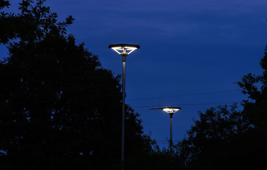 Zwei moderne LED-Straßenlaternen leuchten nachts zwischen Bäumen vor dunkelblauem Himmel.